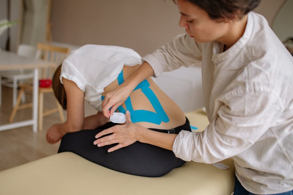 pexels-photo-5794054 A therapist applies blue kinesiology tape to a patient's back in a clinical setting.